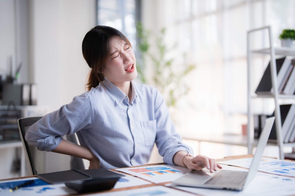 Woman in blue shirt sitting at desk with laptop, holding lower back in pain while working