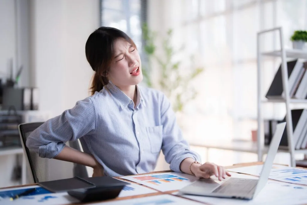 Woman in blue shirt sitting at desk with laptop, holding lower back in pain while working
