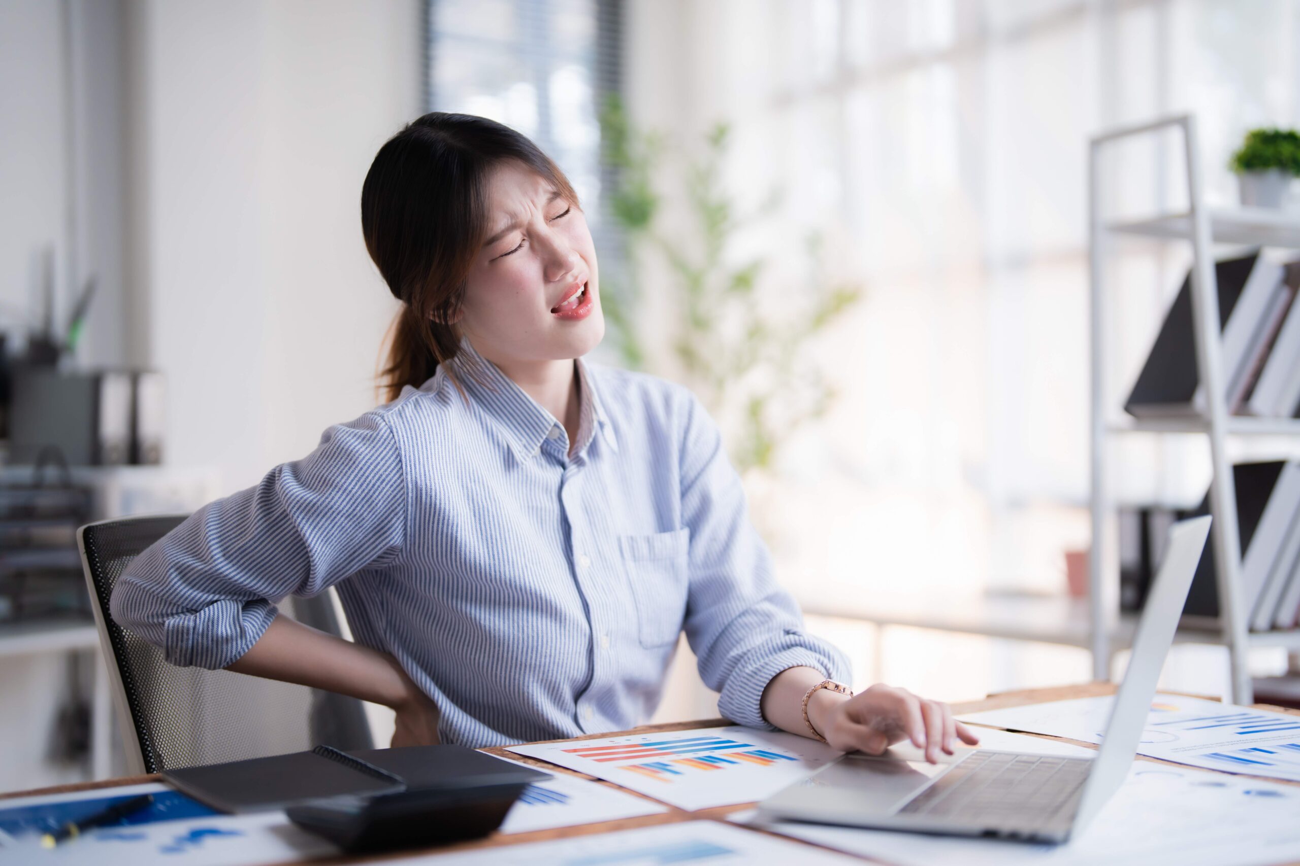 Woman in blue shirt sitting at desk with laptop, holding lower back in pain while working